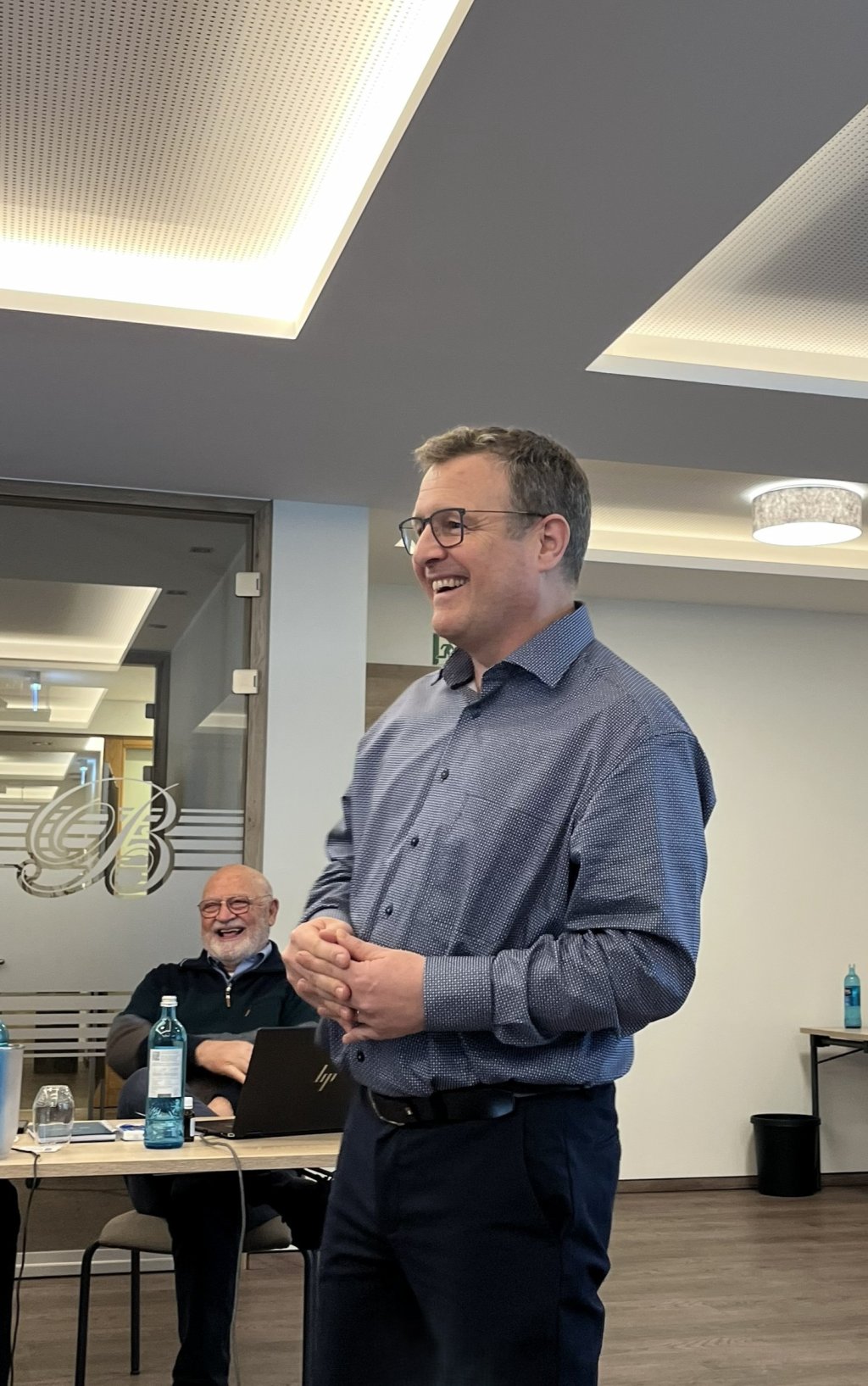 Im Vordergrund l&auml;chelt ein Vortragender w&auml;hrend seiner Ausf&uuml;hrung, ebenso ein Zuh&ouml;rer im Hintergrund.
