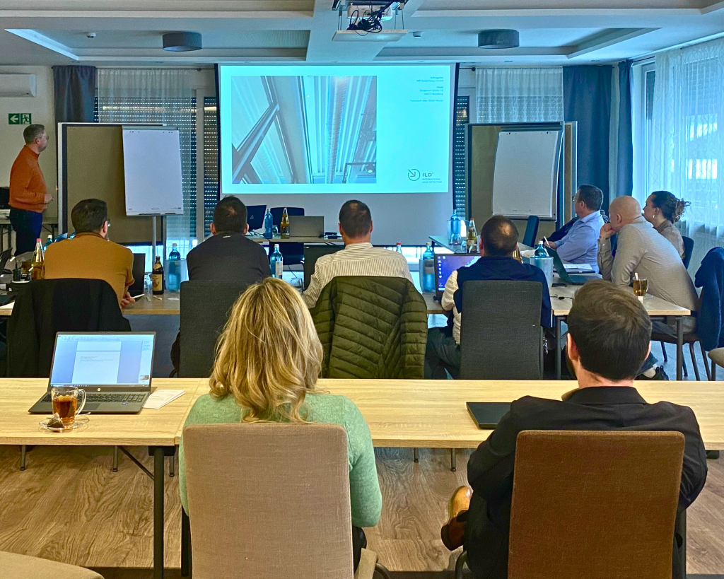 Eine Seminarsituation, Mitarbeiter fotografiert von hinten h&ouml;ren bei einem Vortrag zu, Bilder und Text werden auf eine Leinwand gebeamt.